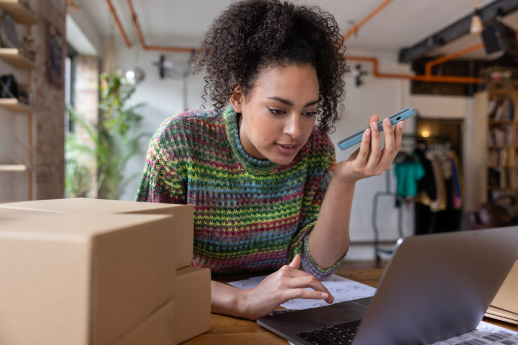 Woman with curly hair in a colorful knit sweater looks at a blue smartphone while seated at a desk with a laptop and cardboard boxes nearby.