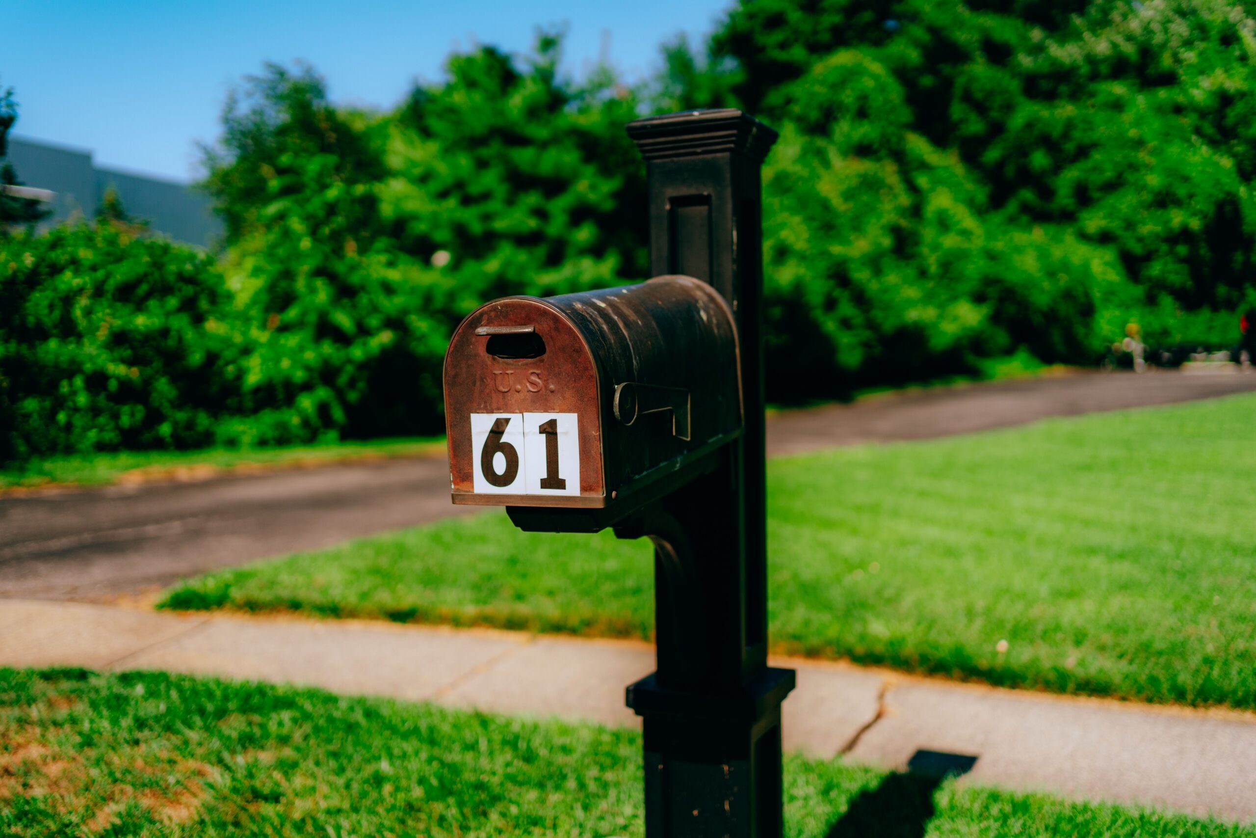 Rusty red mailbox with the number 61 on a black post along a sidewalk, with green lawn and trees in the background.