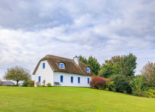 White cottage with a thatched roof on a green lawn, blue window frames and surrounding trees and shrubs.
