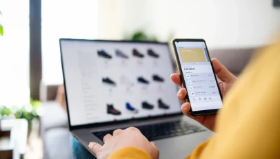 An image depicting a young woman engaging in e-commerce activities using both her smartphone and laptop. She appears focused and is possibly entering or verifying her address details on an online platform. The image illustrates the modern, digital nature of shopping and the importance of accurate address entry in the e-commerce experience.