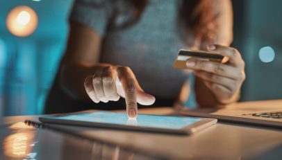 An image displaying an individual using their credit card to make a transaction on an iPad, representing a modern, digital checkout process. This scene captures the essence of streamlined digital transactions, symbolizing the role of advanced technologies like Autoaddress's Address Capture and Autocomplete in facilitating efficient and accurate address entry during online shopping and services.
