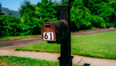 Rusty red mailbox with the number 61 on a black post along a sidewalk, with green lawn and trees in the background.