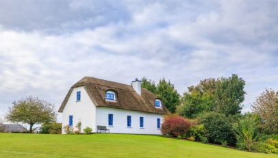 White cottage with a thatched roof and blue-framed windows on a green lawn, with trees in the background.